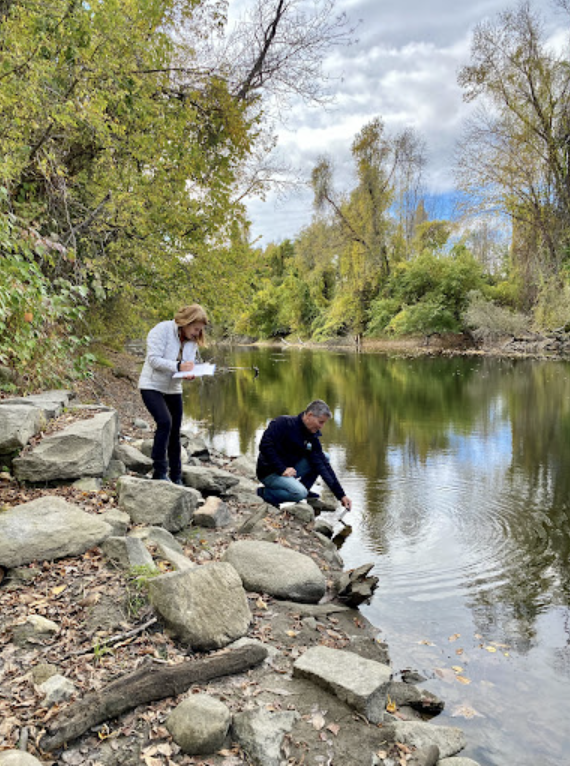 Two people stand on land collecting data from a body of water. 